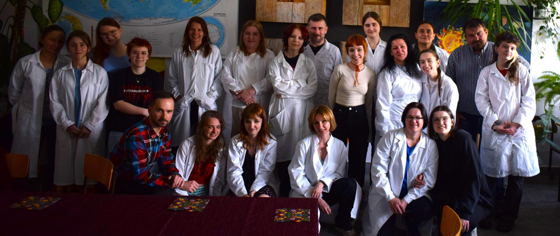 Zdjęcie zbiorowe. Rodzice i uczniowie pozują do zdjęcia w sali lekcyjnej PLSP w Lublinie. W pierwszym szeregu uczniowie i nauczyciel Tomasz Banaszkiewicz są w pozycji przysiadu. W drugim szeregu stoją rodzice i uczniowie. W tle na ścianie wisi geograficzna mapa świata, dwie prace snycerskie obrazujące pory roku. Stoją też duże kwiaty.