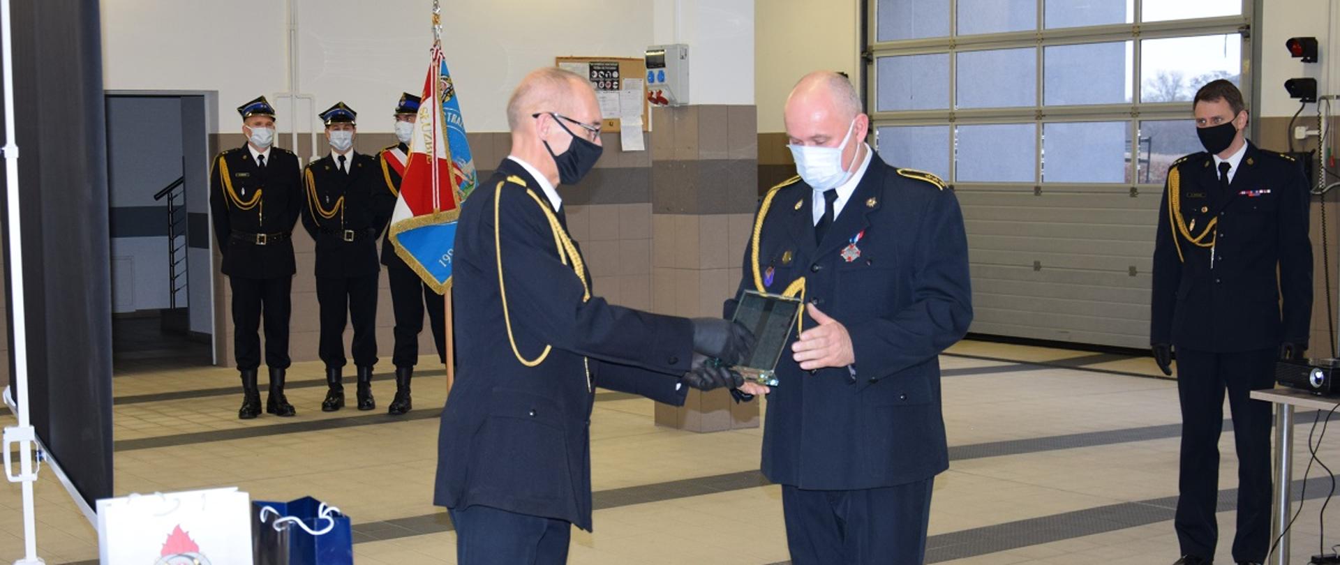Zdjęcie zrobione w garażu bojowym Jednostki Ratowniczo Gaśniczej w Wąbrzeźnie. Na pierwszym planie znajduje się Komendant Powiatowy Państwowej Straży Pożarnej w Wąbrzeźnie starszy brygadier mgr inż. Janusz Woźniak wręczający pamiątkową statuetkę aspirantowi sztabowemu Pawłowi Makowskiemu z okazji przejścia na zaopatrzenie emerytalne. Na drugim planie znajduje się poczet flagowy. Po prawej stronie stoi naczelnik wydziału operacyjno-kontrolno-rozpoznawczego kpt. Robert Stasia.