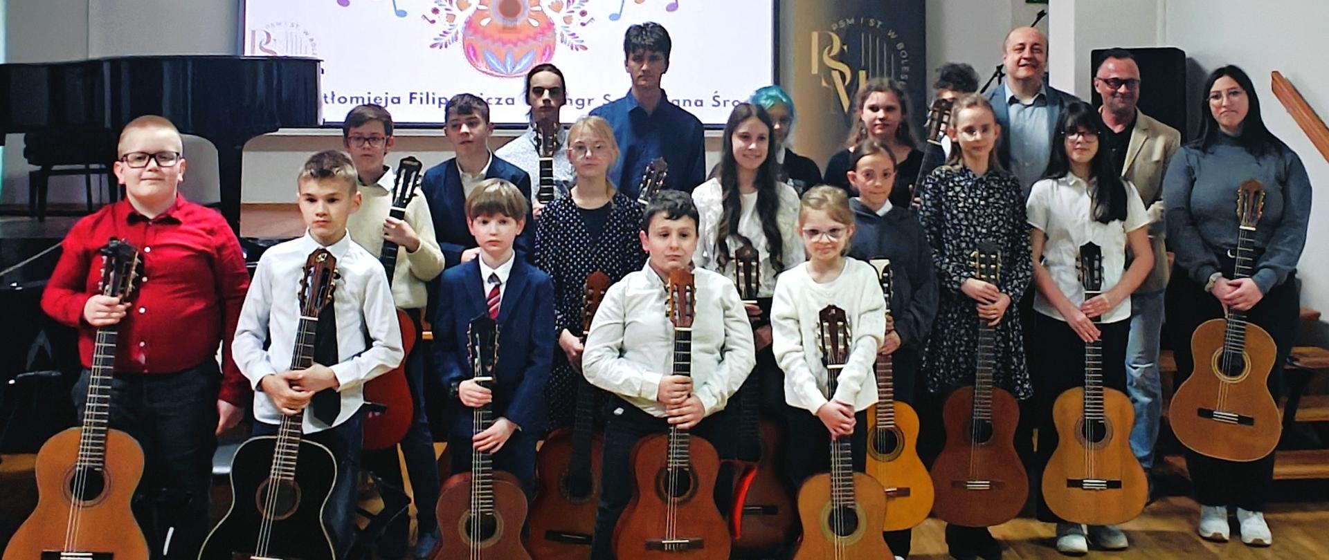 Zdjęcie grupowe, przedstawiające wszystkich wykonawców z gitarą, podczas popisu klasy gitary mgr Bartłomieja Filipowicza i mgr Sebastiana Środy, który odbył się 7 kwietnia 2025 r. w auli szkoły.