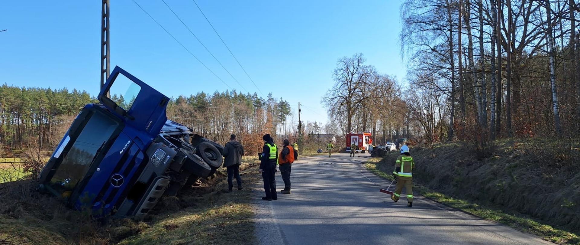 Samochód ciężarowy koloru niebieskiego znajduje się w rowie. Obok stoi policjant w granatowym mundurze i strażak w piaskowym ubraniu specjalnym.