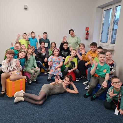 Zdjęcie przedstawia grupę uczniów z klasy I c.6 gr A i I c.4. Dzieci siedzą na pufach lub leżą przed nimi, są ubrane na kolorowo i machają. W tle okno a na podłodze granatowa wykładzina.