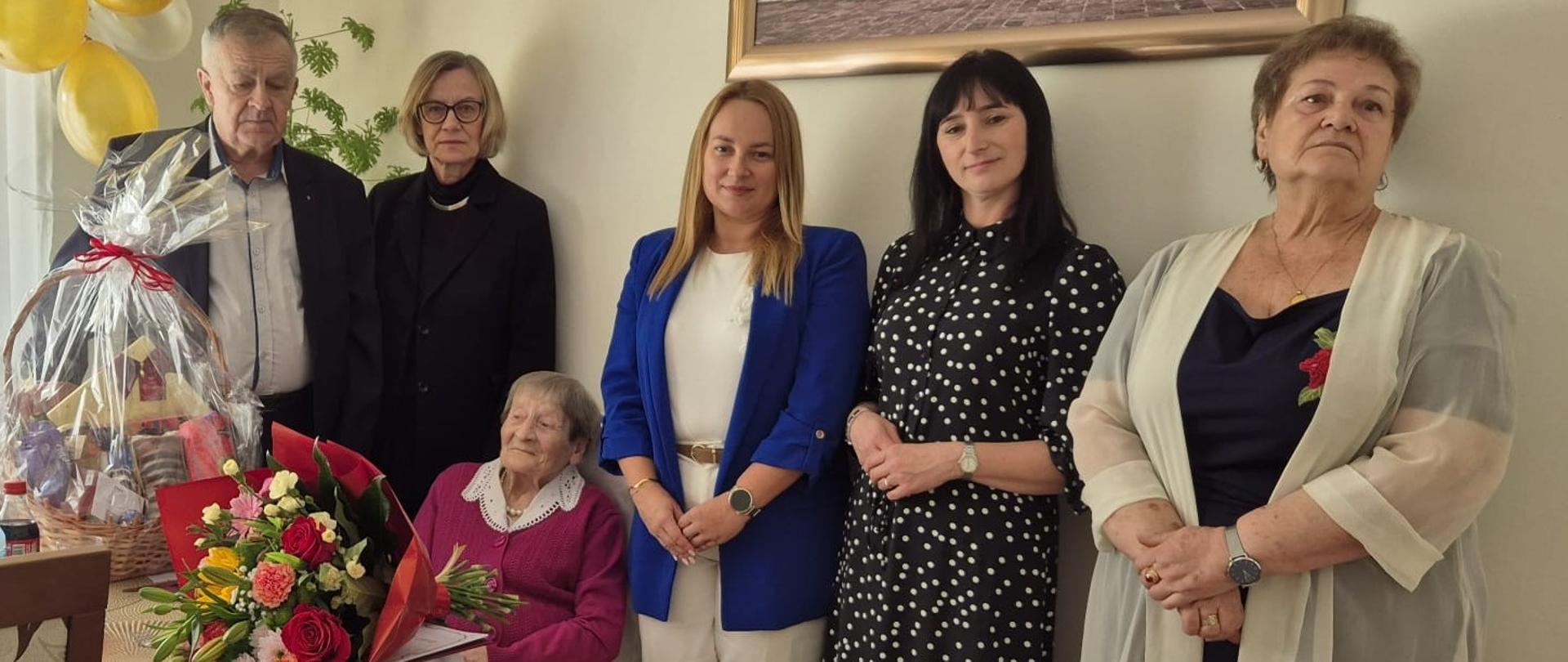 Na fotografii widać grupę stojących osób. W centrum, przy stole z bukietem kwiatów i koszem ze
słodyczami siedzi starsza kobieta – jubilatka obchodząca setne urodziny. Obok niej stoją członkowie
rodziny oraz przedstawiciele lokalnych władz i instytucji.