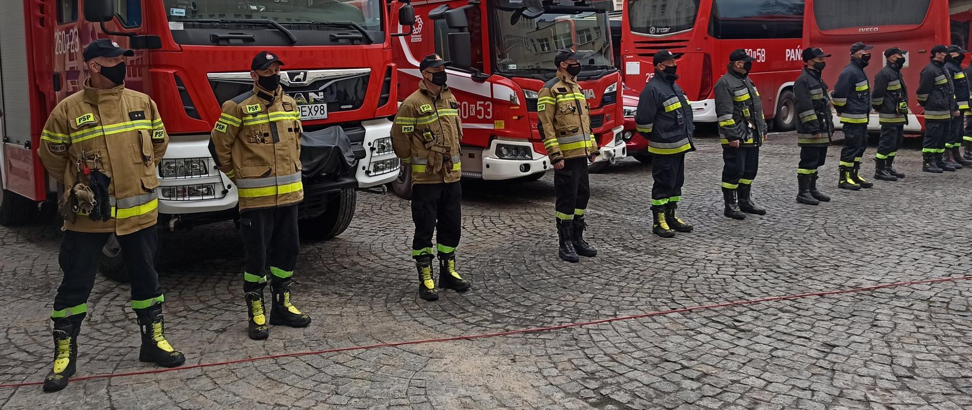 Na zdjęciu strażacy ustawieni przed samochodami pożarniczymi. W tle strażackie samochody ratowniczo-gaśnicze.