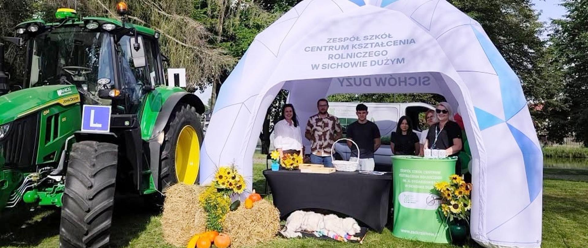 Stoisko wystawiennicze Zespołu Szkół Centrum Kształcenia Rolniczego w Sichowie Dużym podczas dożynek w Rytwianach. Nauczyciele i uczniowie pozują pod białym, dmuchanym namiotem obok zielonego ciągnika rolniczego.