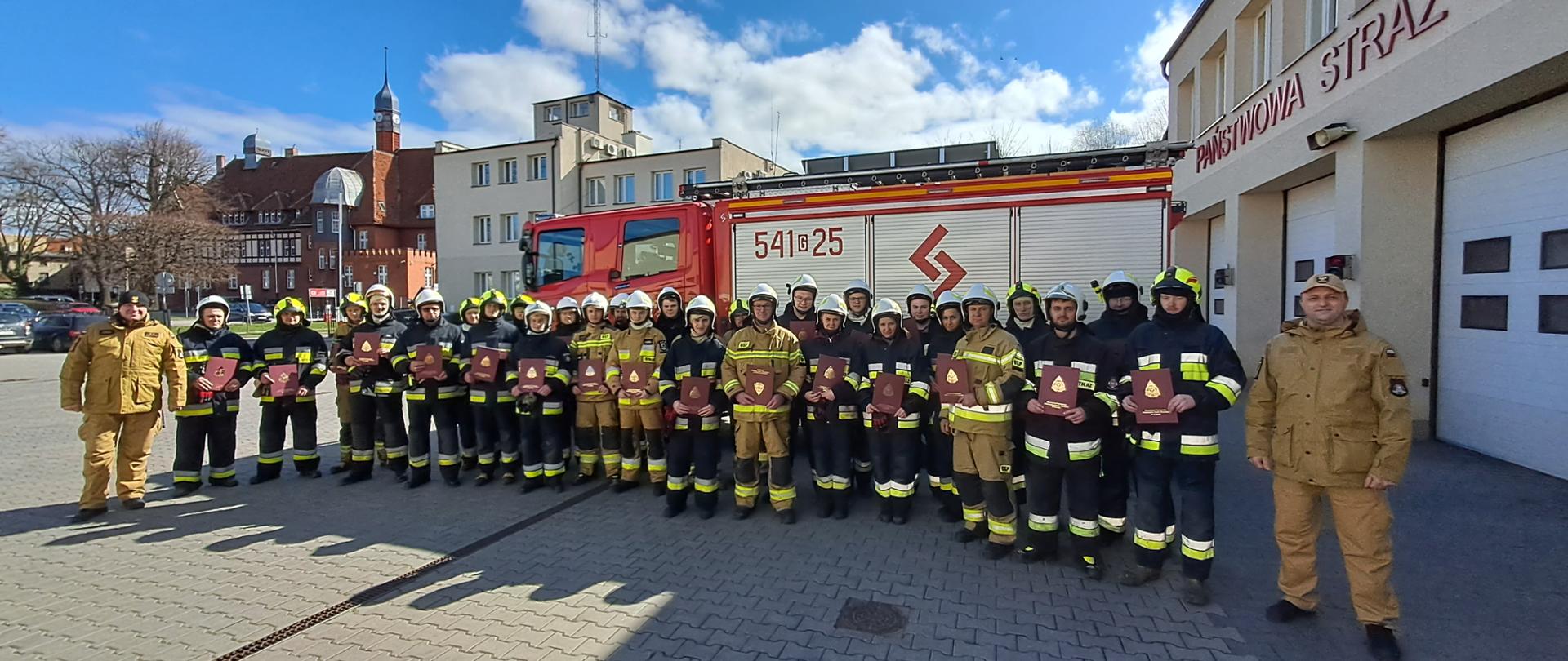 29 absolwentów szkolenia w umundurowaniu specjalnym stoi na grupowym zdjęciu z dwoma instruktorami szkolenia. Z tyłu ciężki samochód ratowniczo-gaśniczy, w tle budynek Komendy i ratusz miejski.
