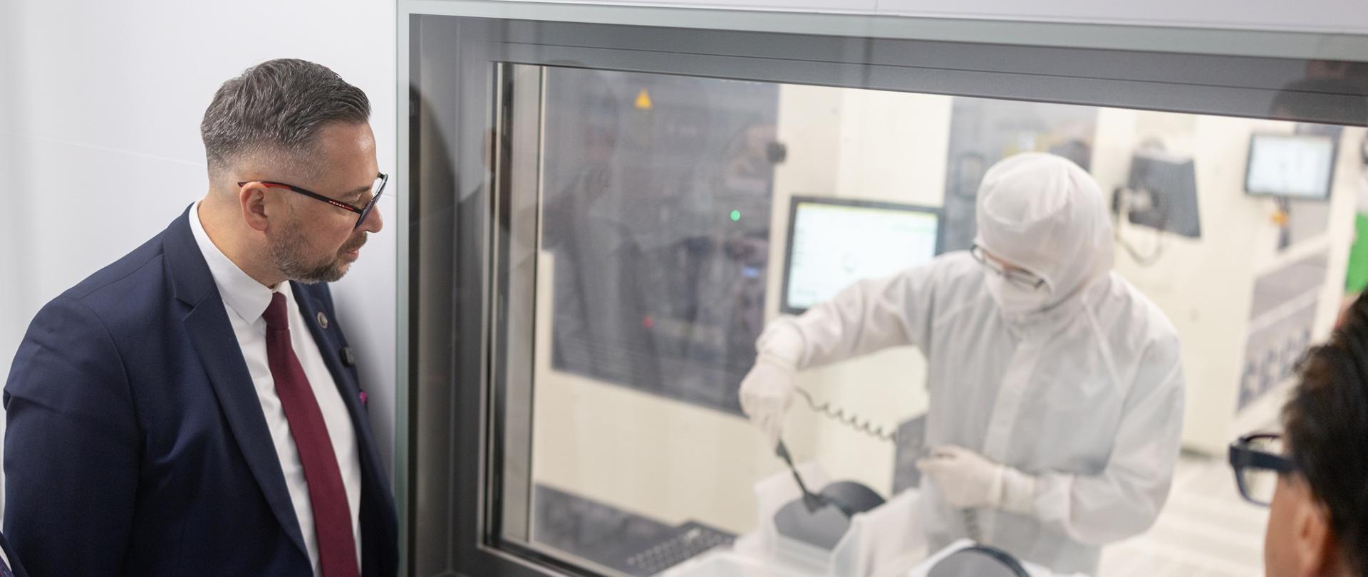 Minister Kulasek stoi przy białej ścianie, patrzy przez oszklone okno za którym widać postać w białym fartuchu obsługującą urządzenie.