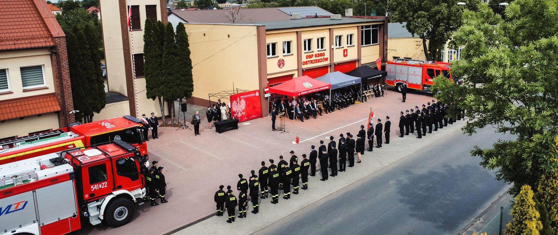Zdjęcie z lotu ptaka placu na którym odbywają się obchody Dnia Strażaka. Na pierwszym planie strażacy w pododdziale obok wozów strażackich. Na drugim planie namioty z gośćmi i budynek remizy OSP.