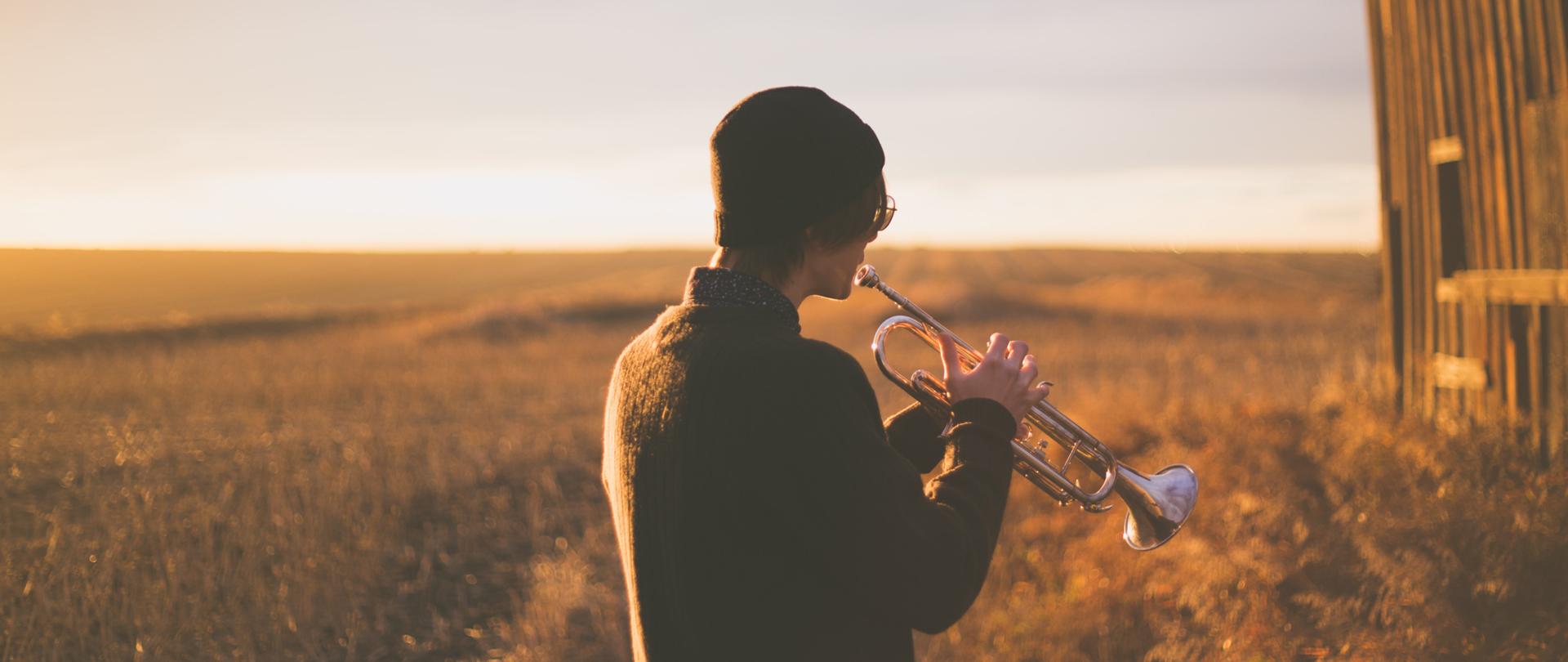 Fotografia przedstawia młodego mężczyznę ubranego w ciemną, wełnianą czapkę oraz sweter, stojącego na łące i grającego na trąbce, mężczyzna odwrócony jest plecami, po prawej stronie zdjęcia widać fragment drewnianej szopy