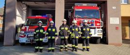 Widok z przodu. Pięciu strażaków OSP Gierałtowice stoi w ubraniach specjalnych przed samochodami pożarniczym przed garażem OSP. 