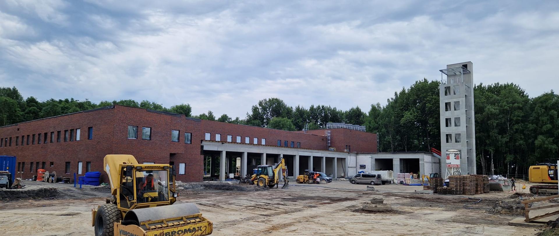 Na zdjęciu widoczny plac budowy na terenie nowo budowanej komendy miejskiej Państwowej Straży Pożarnej w Rudzie Śląskiej z "lotu ptaka". Na zdjęciu po lewej stronie widać budowaną siedzibę komendy, składająca się z parteru z garażami i piętra z cegły. Na placu stoją samochody i sprzęt budowlany - koparka i walec. W tle za ogrodzeniem drzewa.