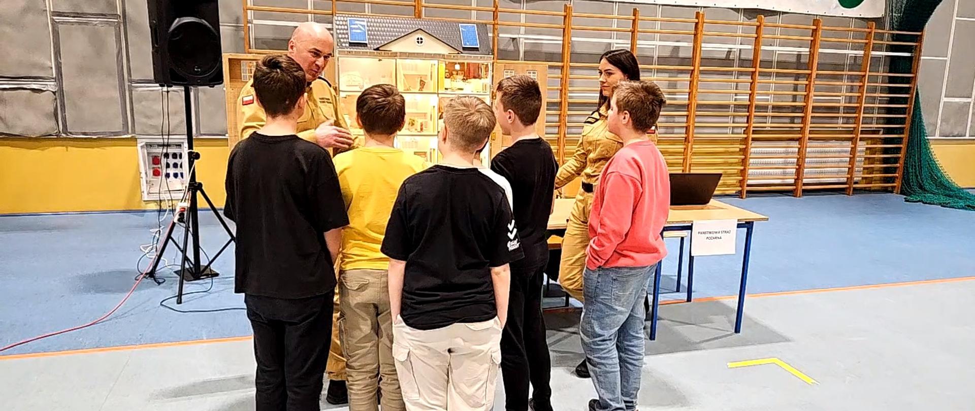 Zdjęcie przedstawia strażaków podczas targów edukacyjnych. Strażacy przy pomocy mobilnego symulatora zagrożeń pożarowych omawiają zagrożenia. 