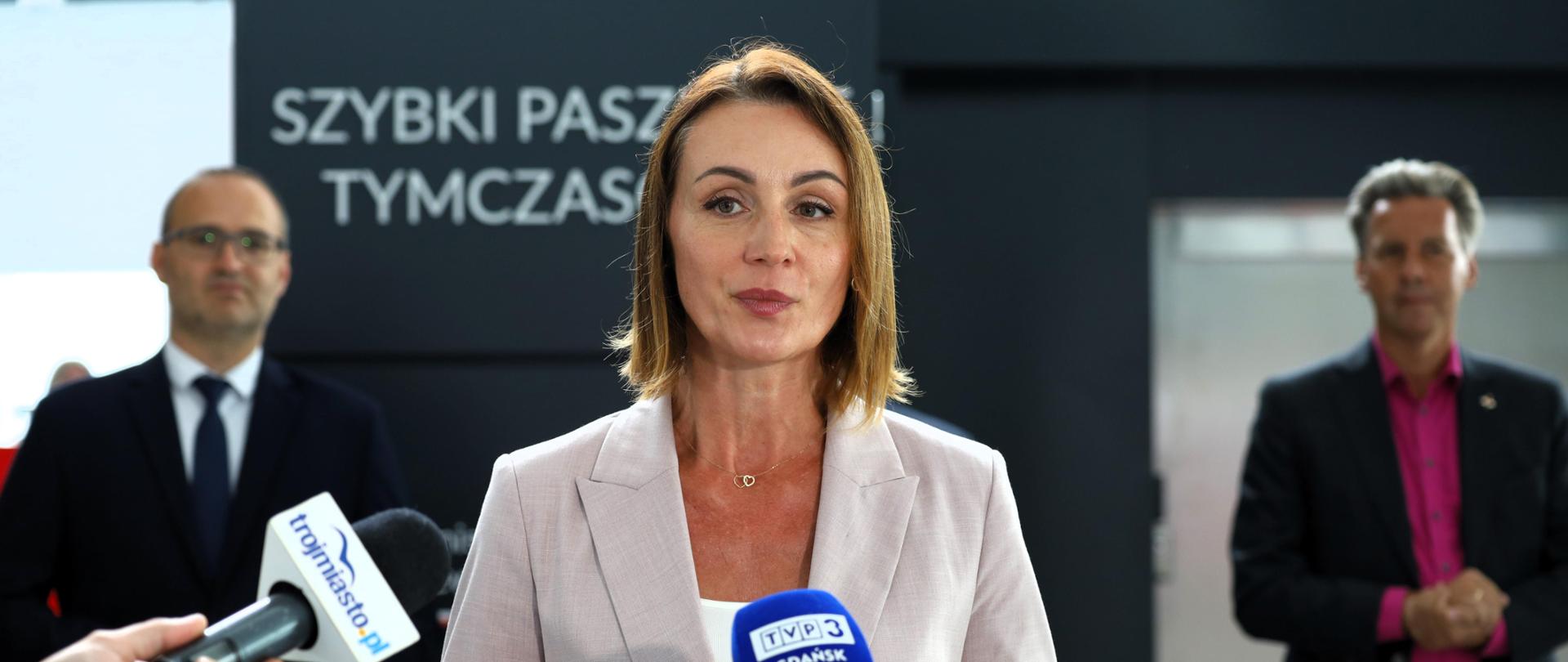 Kobieta (wojewoda) stoi przed mikrofonami. W tle napis Szybki Paszport Tymczasowy