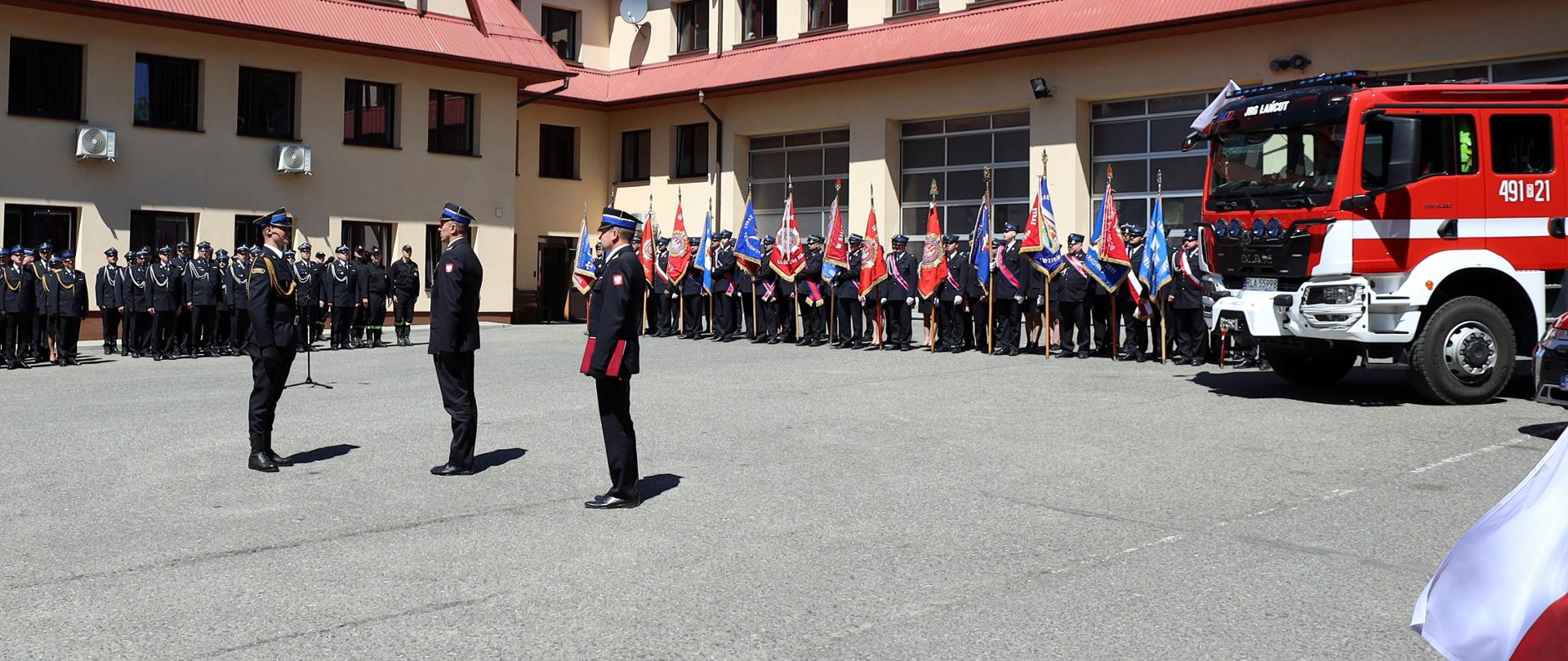 Zdjęcie zrobione na zewnątrz, na placu wewnętrznym Komendy Powiatowej Państwowej Straży Pożarnej. Na zdjęciu strażacy ustawieni na uroczystej zbiórce. Na pierwszym planie dowódca uroczystości składa meldunek oficerowi Państwowej Straży Pożarnej. W tle widać strażaków uczestniczących w uroczystości oraz szereg strażaków trzymających sztandary jednostek. Po prawej stronie widać przód samochodu pożarniczego.