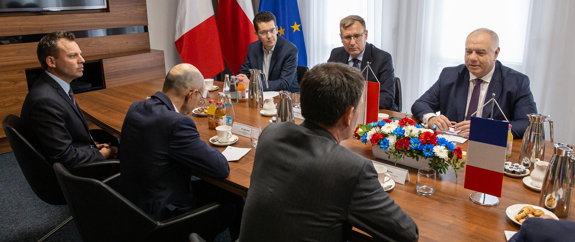 Na zdjęciu z lewej strony siedzą ambasador Francji Frederic Billet i przedstawiciele firmy Total Energies, z prawej strony wicepremier Jacek Sasin oraz wiceminister Maciej Małecki. Na stole proporczyki i kwiaty. 
