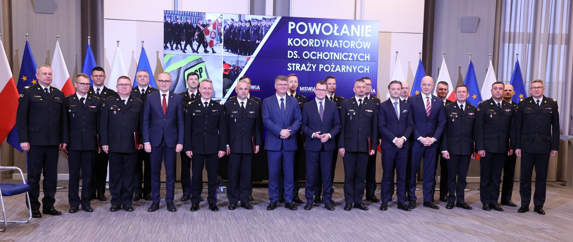 na zdjęciu grupa strażaków w mundurach wyjściowych, koordynatorów do spraw ochotniczych straży pożarnych i komendantów głównych psp oraz kilku mężczyzn w garniturach , stojących frontem do fotografa w tle ekran z wyświetlonymi zdjęciami oraz napisem powołanie koordynatorów ds. ochotniczych straży pożarnych oraz flagi polski i unii europejskiej