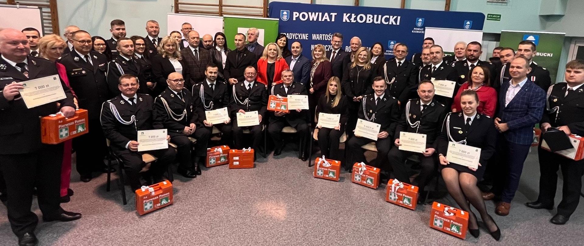 Grupa kilkudziesięciu osób pozuje do zdjęcia. Część z nich stoi w dwóch rzędach a część siedzi na krzesłach przed nimi. Większość osób jest w mundurach OSP i trzyma w rękach promesy z kwotami dofinansowania. Przed nimi stoją małe skrzynki z apteczkami. Z tyłu granatowa ścianka powiatu kłobuckiego. i rollupy z logo KRUS.