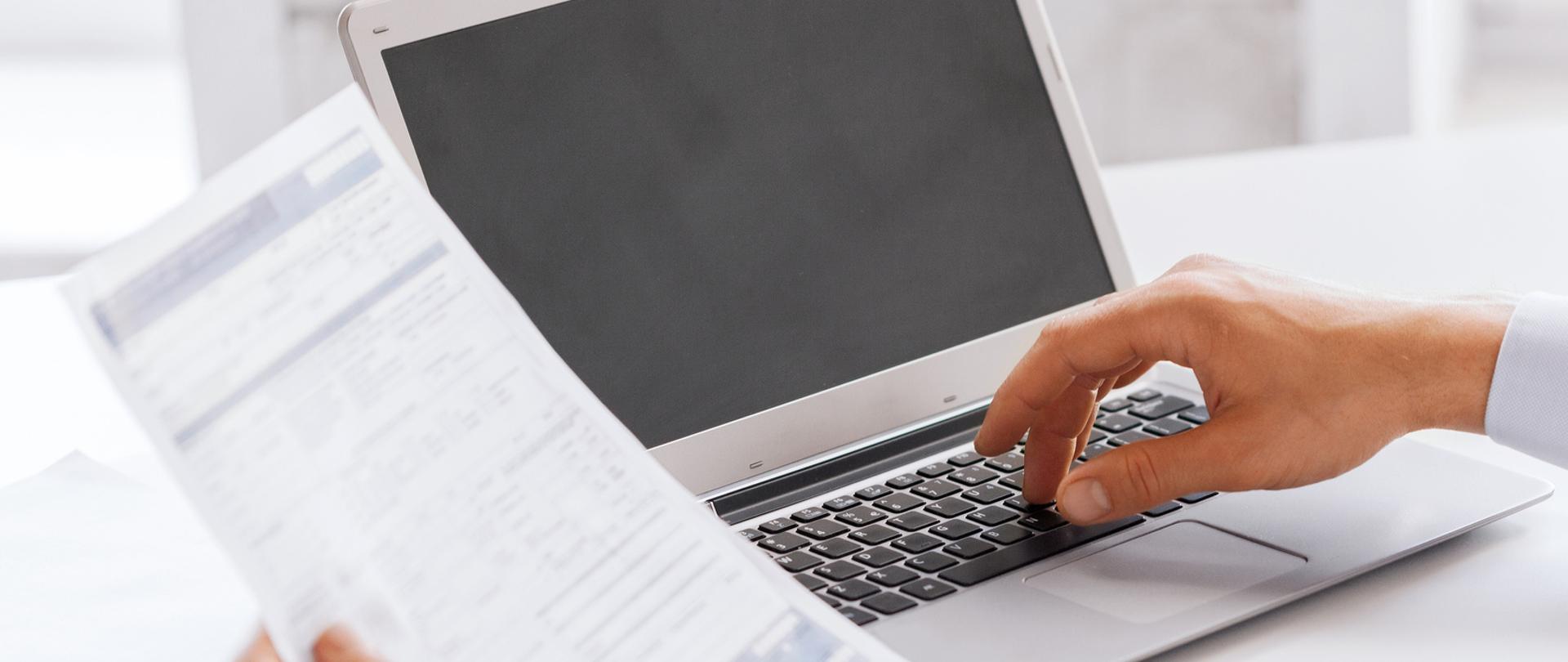Mężczyzna przed laptopem, jedną rękę trzyma na klawiaturze, w drugiej ręce ma dokument