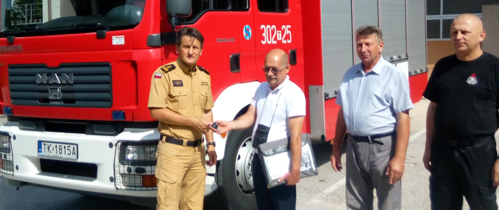 Zdjęcie przedstawia Komendanta Miejskiego Państwowej Straży Pożarnej w Kielcach przekazującego kluczyki do samochodu pożarniczego, który stanowi tło .
