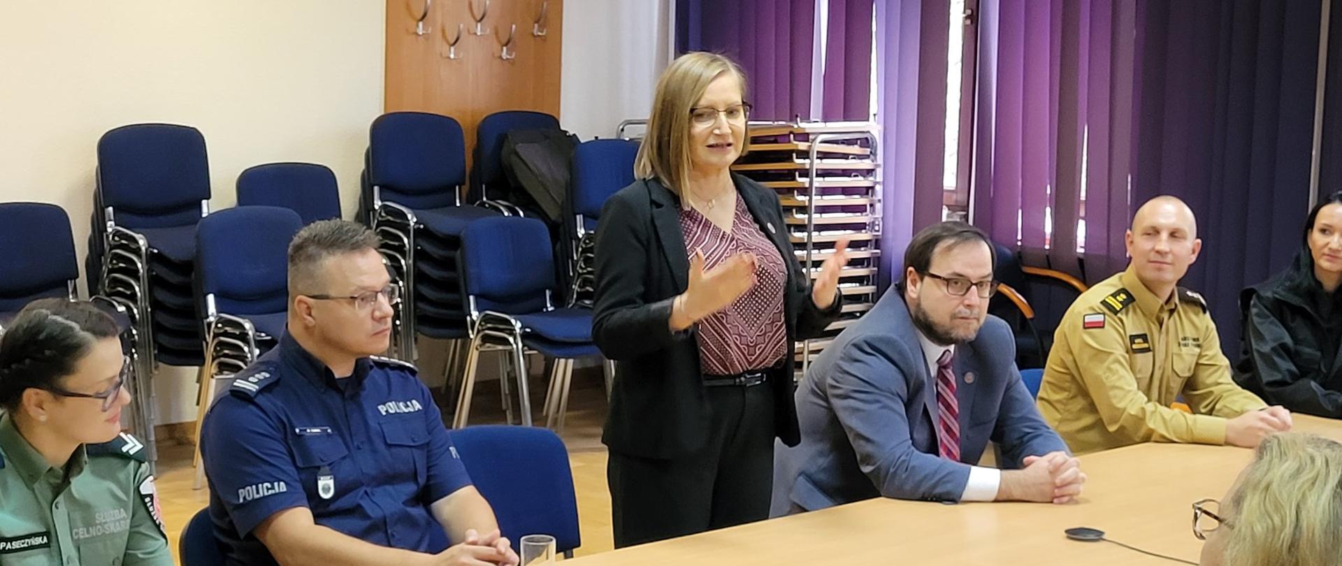 Kobieta stoi przy stole, Przemawia i gestykuluje. Obok niej siedzą ludzie, cześć z nich jest ubrana w mundury służbowe.