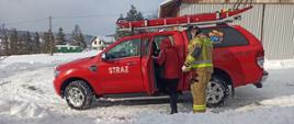 Na zdjęciu widoczny strażak na tle samochodu strażackiego do którego wsiada kobieta w celu przetransportowania jej do punktu szczepień.