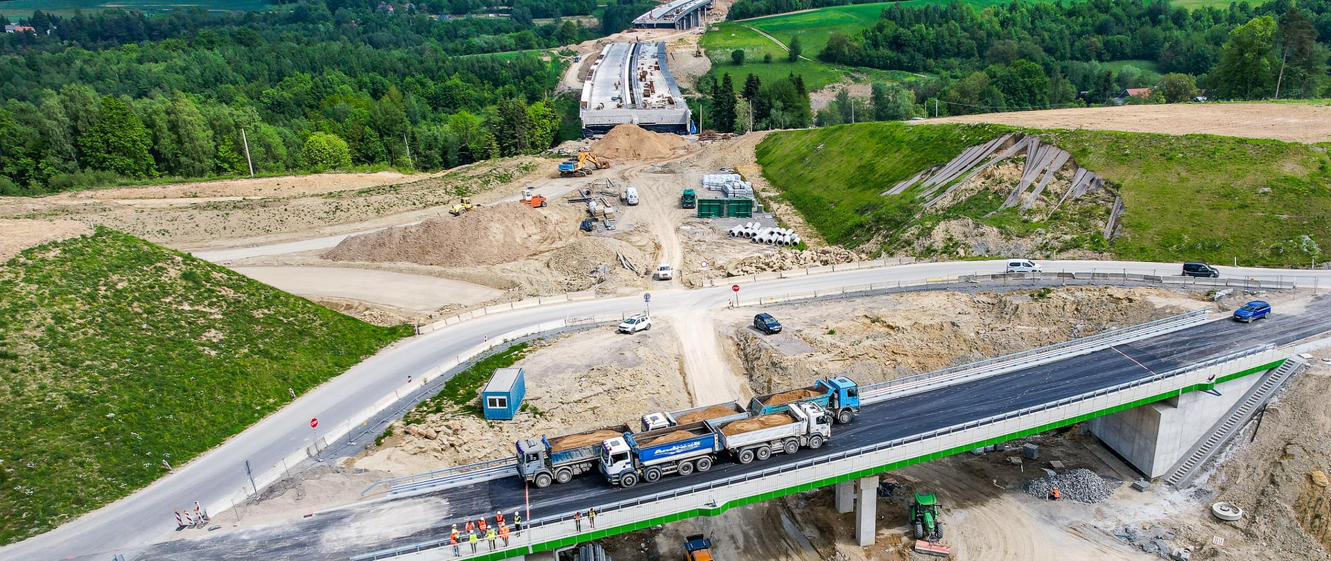 Plac budowy drogi S19 Babica - Jawornik - nowy wiadukt nad wykopem, na którym stoi 5 ciężarówek z piaskiem. Wokół pracują koparki i inne maszyny, a obok stoją robotnicy w kaskach; w tle widać zielone wzgórza i las. Zdjęcie lotnicze.