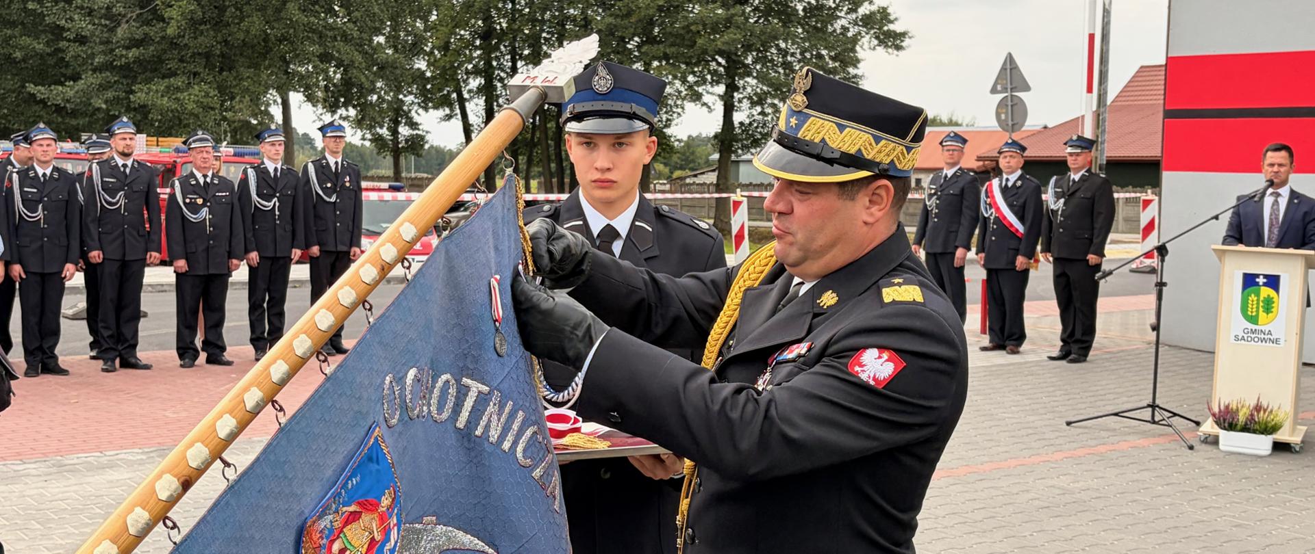 „Ceremonia dekoracji sztandaru jednostki OSP Morzyczyn-Włóki. Komendant Wojewódzki PSP nadbryg. Artur Gonera w asyście strażaka przypina odznaczenie do płata sztandaru. W tle zgromadzeni strażacy i zaproszeni goście.”