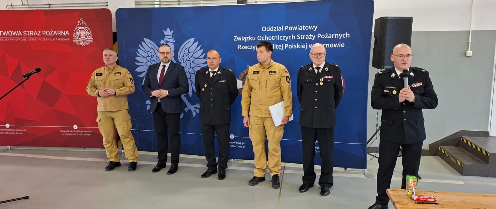 Strażacy oraz zaproszony gość stoją przed bilbordami w jednym szeregu. Strażacy ubrani są w piaskowe mundury oraz galowe mundury, zaproszony gość ubrany jest w garnitur