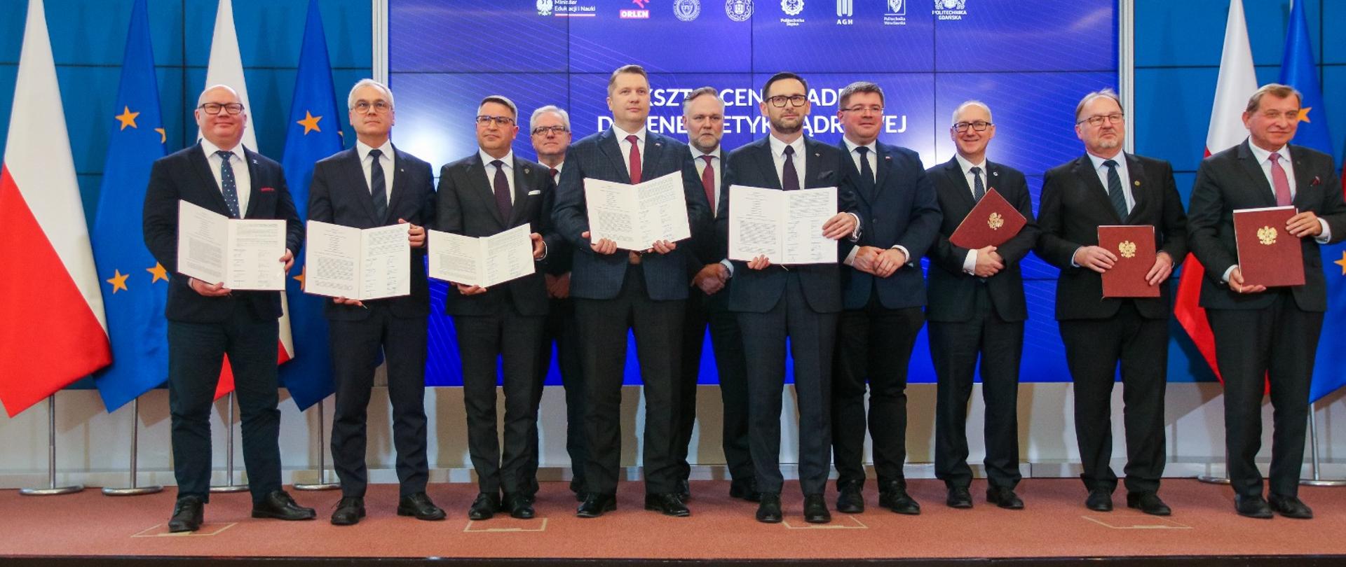 Grupa mężczyzn w garniturach stoi w rzędzie przed niebieską ścianą, trzymają w rękach dokumenty, za nimi z lewej flagi Polski i UE.