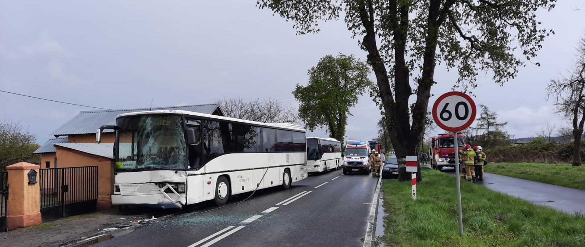 Na zdjęciu 2 białe autokary po kolizji, karetka, samóchod pożarniczy oraz strażacy w umundurowaniu bojowym wykonujący czynności.