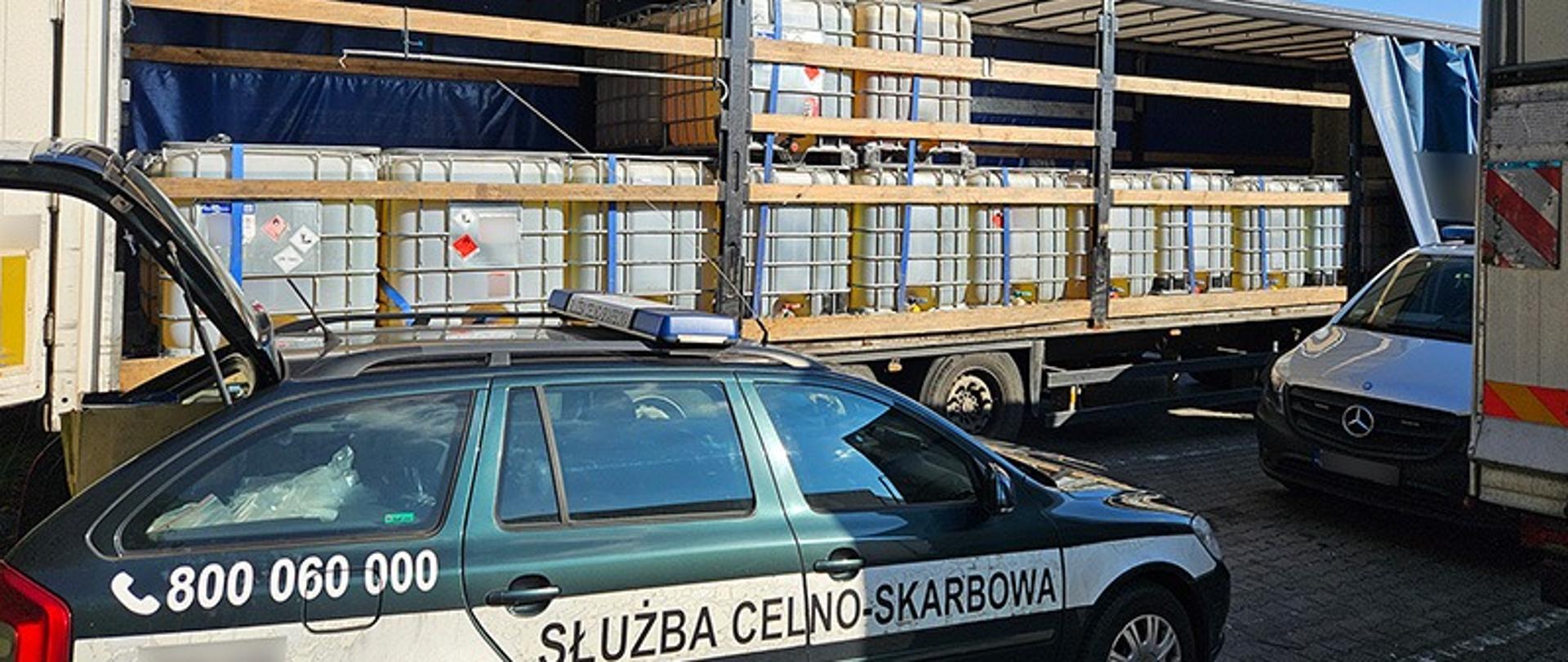 Zdjęcie przedstawia kontrolę transportu drogowego prowadzoną przez Służbę Celno-Skarbową. Na pierwszym planie widoczny jest tył i bok oznakowanego, ciemnozielonego radiowozu z napisem „SŁUŻBA CELNO-SKARBOWA” oraz numerem telefonu „800 060 000”. W tle znajduje się naczepa ciężarówki z odsłoniętą plandeką, wypełniona piętrowo ułożonymi zbiornikami typu IBC (mauzery). Zbiorniki są zabezpieczone pasami transportowymi i posiadają etykiety ostrzegawcze dotyczące substancji chemicznych. W górnych rogach zdjęcia widnieją stałe elementy graficzne: logotyp KAS (Krajowa Administracja Skarbowa) po lewej stronie, adres strony gov.pl/kas po prawej stronie.
