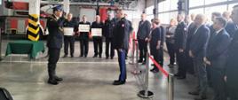Zdjęcie przedstawia dowódcę uroczystości składającego meldunek Wielkopolskiego Komendantowi Wojewódzkiemu PSP. Dowódca oddaje honor poprzez salutowanie. W tle stoją druhowie OSP, którzy trzymają w rękach promesy. Z prawej strony stoją zaproszeni goście.