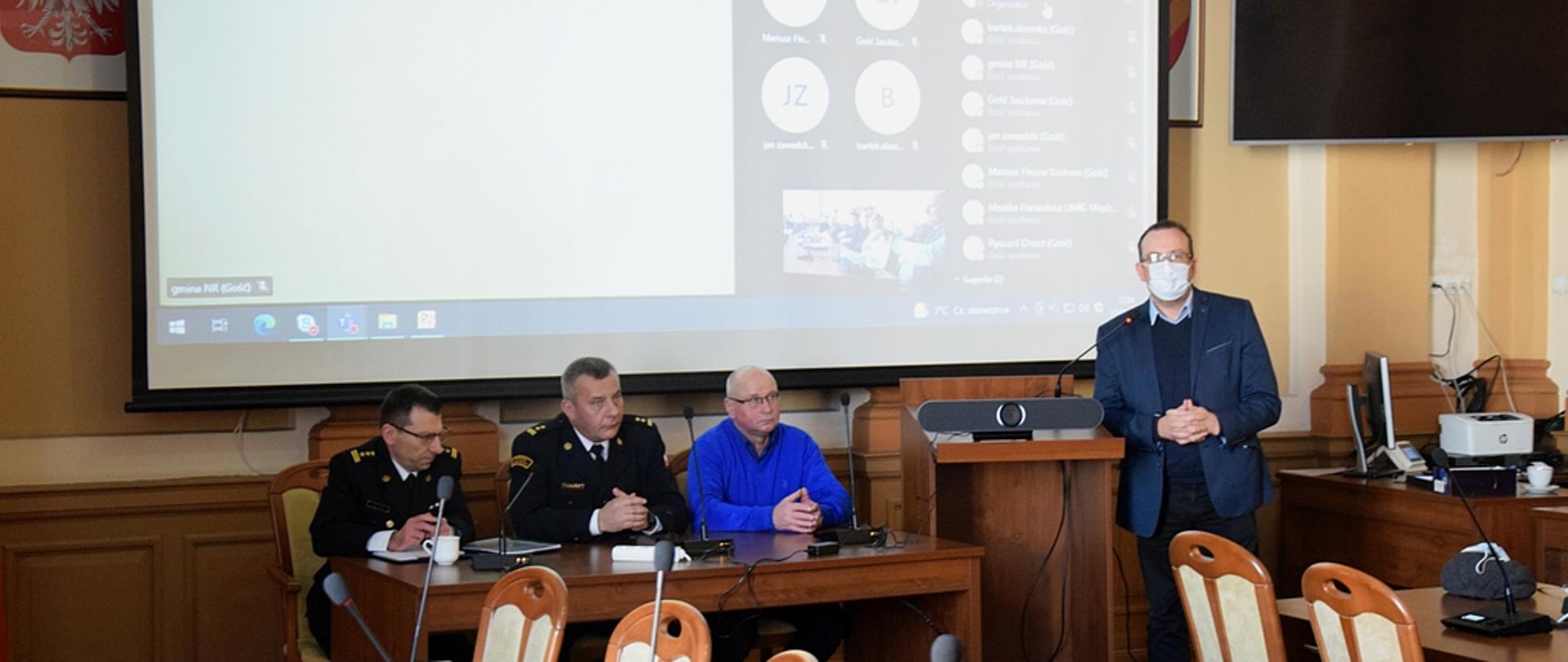 Widok na salę konferencyjną. Po środku przy stole siedzą od lewej: Zastępca Komendanta Powiatowego PSP w Kłodzku , Komendant Powiatowy PSP w Kłodzku oraz Prezes Zarządu Oddziału Powiatowego Związku Ochotniczej straży Pożarnej Rzeczypospolitej Polskiej w Kłodzku. Za nimi na ścianie rozwinięty ekran multimedialny. Po prawej stronie stołu stoi Starosta Powiatu Kłodzkiego. 