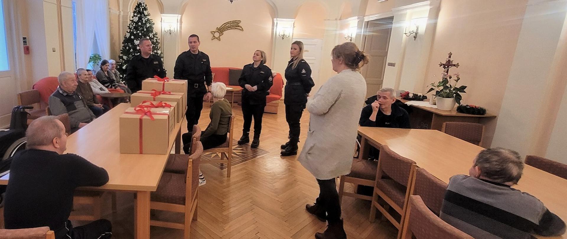 1. Grupa ludzi znajduje się w pomieszczeniu. Na środku stoi czterech strażaków. Strażacy są ubrani w czarne mundury dowódczo-sztabowe. Obok strażaków stoi pani w szarym swetrze. W lewym rogu stoi choinka. W tle na ścianie wisi gwiazda. Po prawej stronie na stoliku stoi kwiat i krzyż. Po prawej i lewej stronie znajdują się stoły. Przy stołach siedzą mieszkańcy Domu Pomocy Społecznej. 