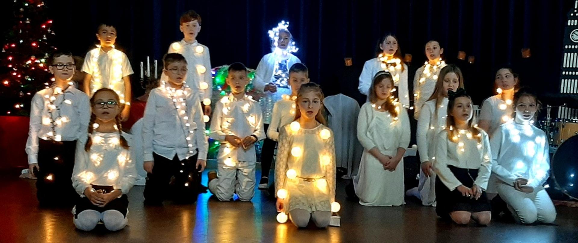 Zdjęcie wykonane w sali koncertowej szkoły na tle scenografii "Opowieść wigilijna". Uczniowie ubrani w stroje białe lub biało-czarne, przyozdobieni światełkami.
