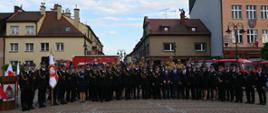 Zgromadzeni uczestnicy apelu pozują do zdjęcia. W lewej części zdjęcia poczet sztandarowy. W tle budynki żorskiej starówki. 