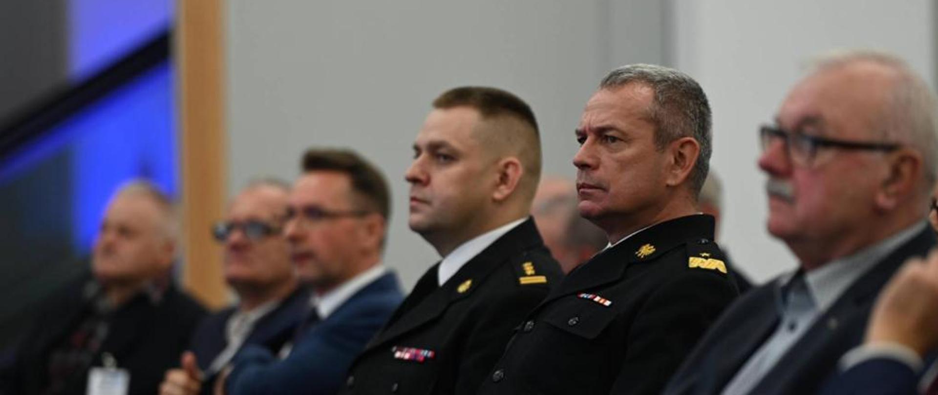 Zastępca Komendanta Głównego PSP nadbryg. Arkadiusz Przybyła podczas konferencji tematycznej podczas odbywających się w Poznaniu Międzynarodowych Targów Ochrony Środowiska POLECO 2021. Obok komendant siedzi bryg. Sławomir Brant rzecznik prasowy Wielkopolskiego Komendanta Wojewódzkiego Państwowej Straży Pożarnej. W tle pozostali uczestnicy konferencji. 