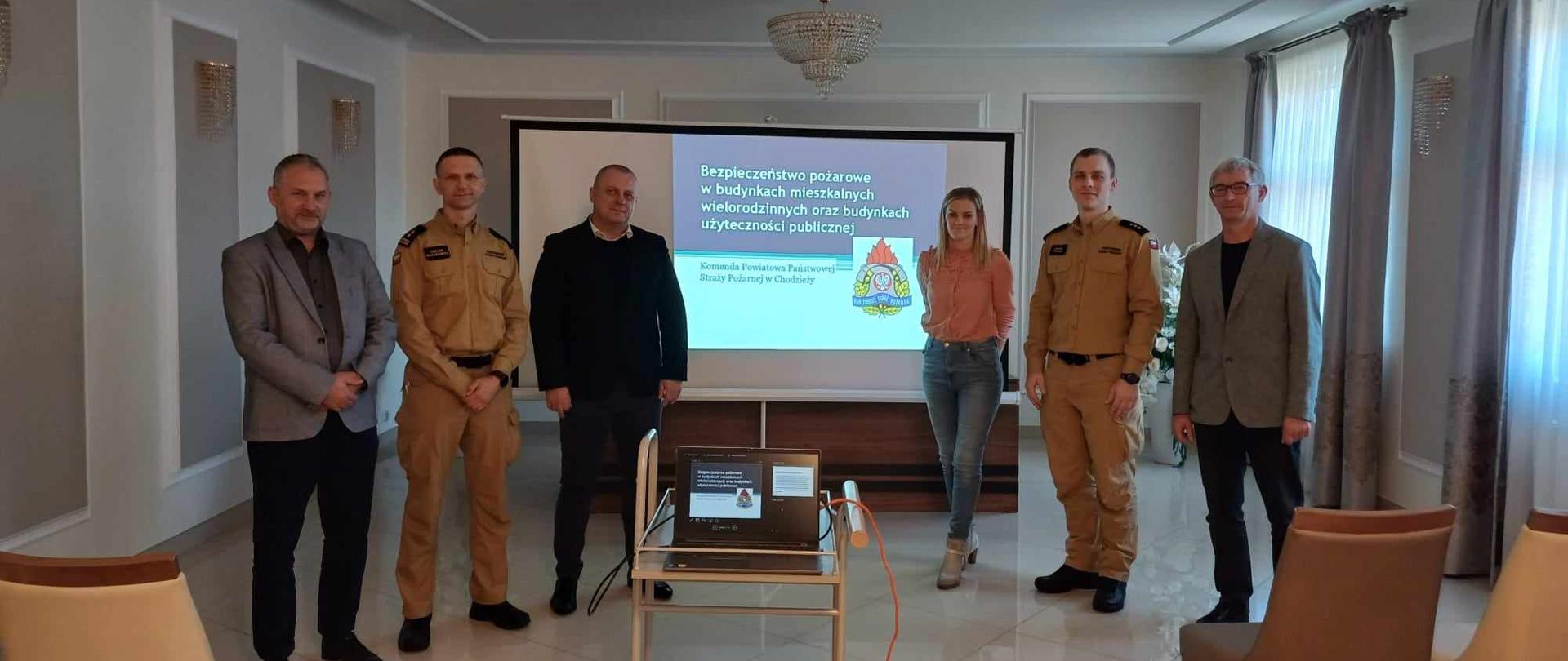 Zdjęcie przedstawia strażaków i przedstawicieli zarządców budynków, którzy wzięli udział w spotkaniu i szkoleniu dotyczącym bezpieczeństwa pożarowego w budynkach. .
W tle pomieszczenie.
