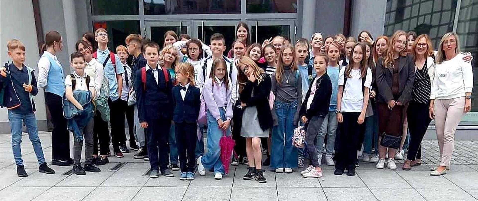 Zjecie przedstawie grupe uczniów wraz z nauczycielami podczas wycieczki do filharmonii zielonogórskiej. W tle budynek filharmonii