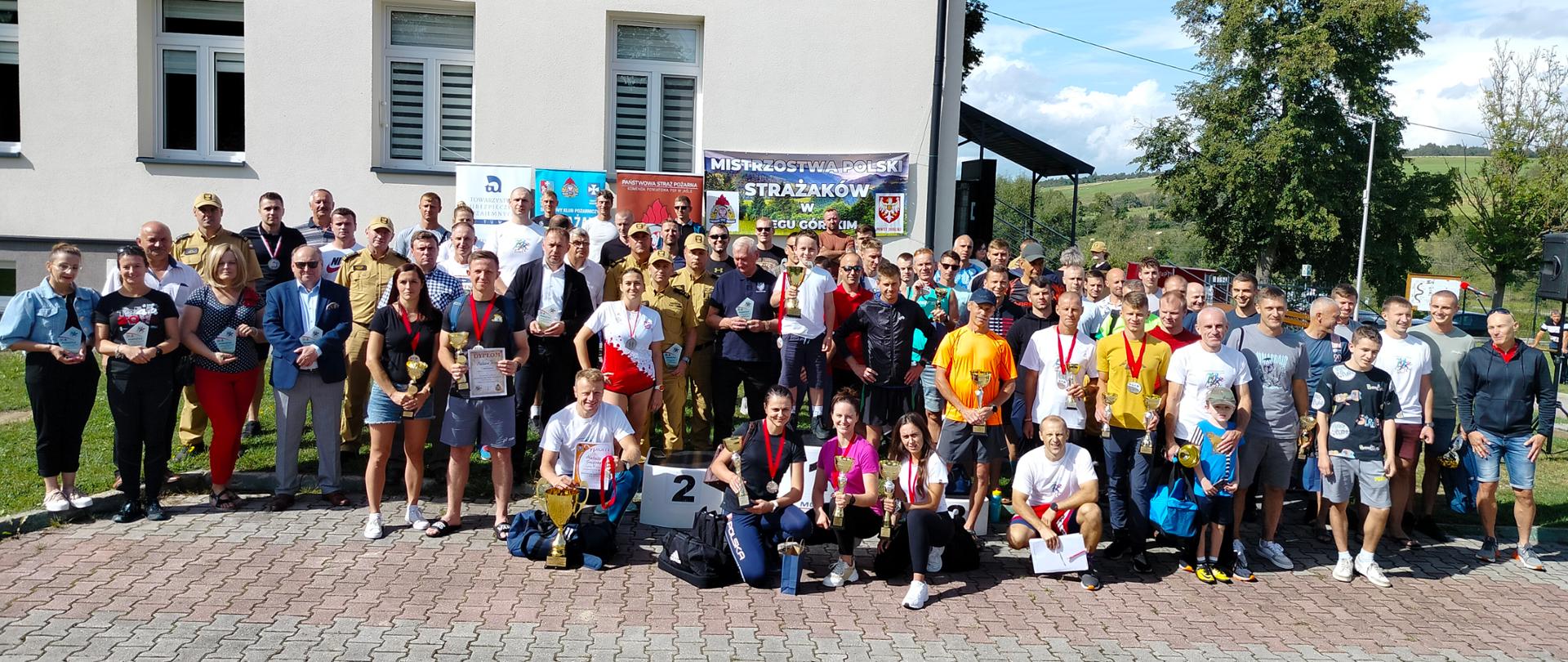 Grupowe zdjęcie zawodników, organizatorów. Osoby cywilne i strażacy w mundurach, zawodnicy trzymają w dłoniach puchary, dyplomy a na szyjach mają zawieszone medale