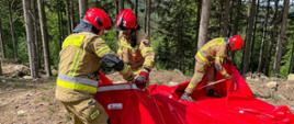 Na zdjęciu widać trzech strażaków w umundurowaniu musztardowym oraz bojowym Strażacy rozkładają zbiornik brezentowy koloru czerwonego. W tle widać drzewa. Zdjęcie wykonane w lesie na drodze. 