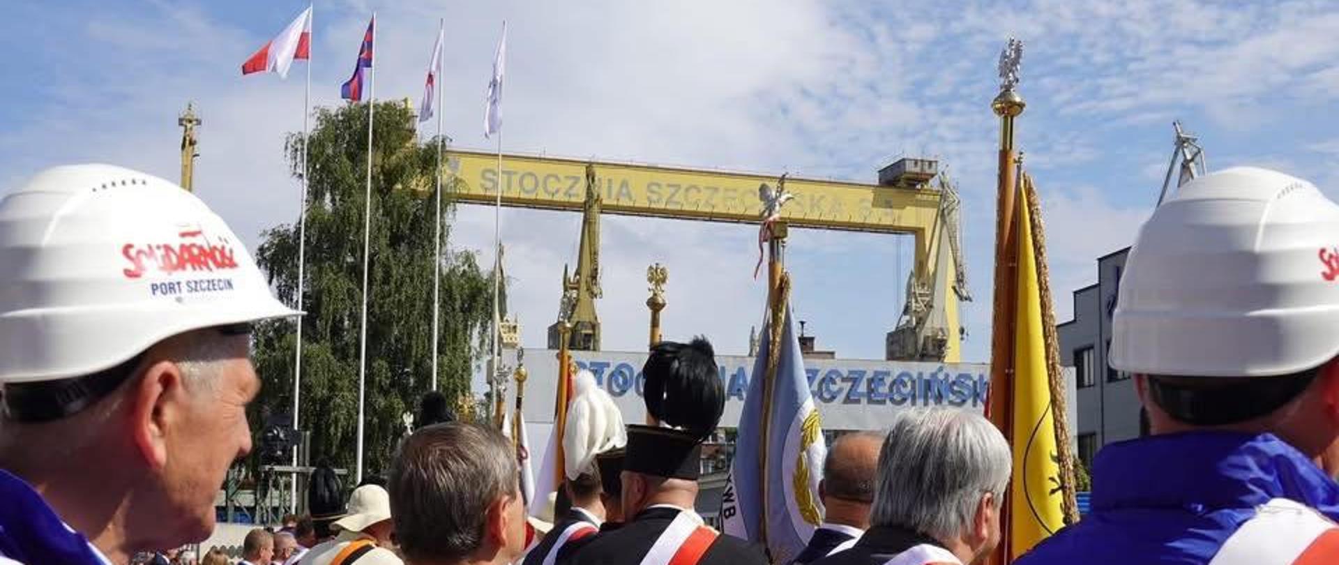 
Zdjęcie przedstawia dużą grupę osób, które uczestniczą w ceremonii na zewnątrz, prawdopodobnie w stoczni. Większość osób jest zwrócona plecami do aparatu, ale można zobaczyć, że niektórzy mają na sobie białe kaski ochronne z czerwonymi napisami, prawdopodobnie powiązanymi z ruchem Solidarność.
Z przodu, po lewej stronie, widać mężczyznę w białym kasku, ubranego w niebieską kurtkę ze szkarłatną szarfą. W jego tle i po prawej stronie, widać innych ludzi w podobnym stroju. Niektórzy z nich, szczególnie ci w środku, są ubrani w oficjalne, czarne stroje z białymi pióropuszami na głowach i szarfami w narodowych barwach.
W tle, za grupą, widać ogromny, żółty dźwig portowy z czarnym napisem „Stocznia Szczecińska”. Na zdjęciu widoczne są też liczne flagi – zarówno polskie, jak i te związane z Solidarnością, umieszczone na masztach, które powiewają na wietrze. Niebo w tle jest czyste z kilkoma białymi chmurami. Słońce mocno oświetla scenę, rzucając cienie.
