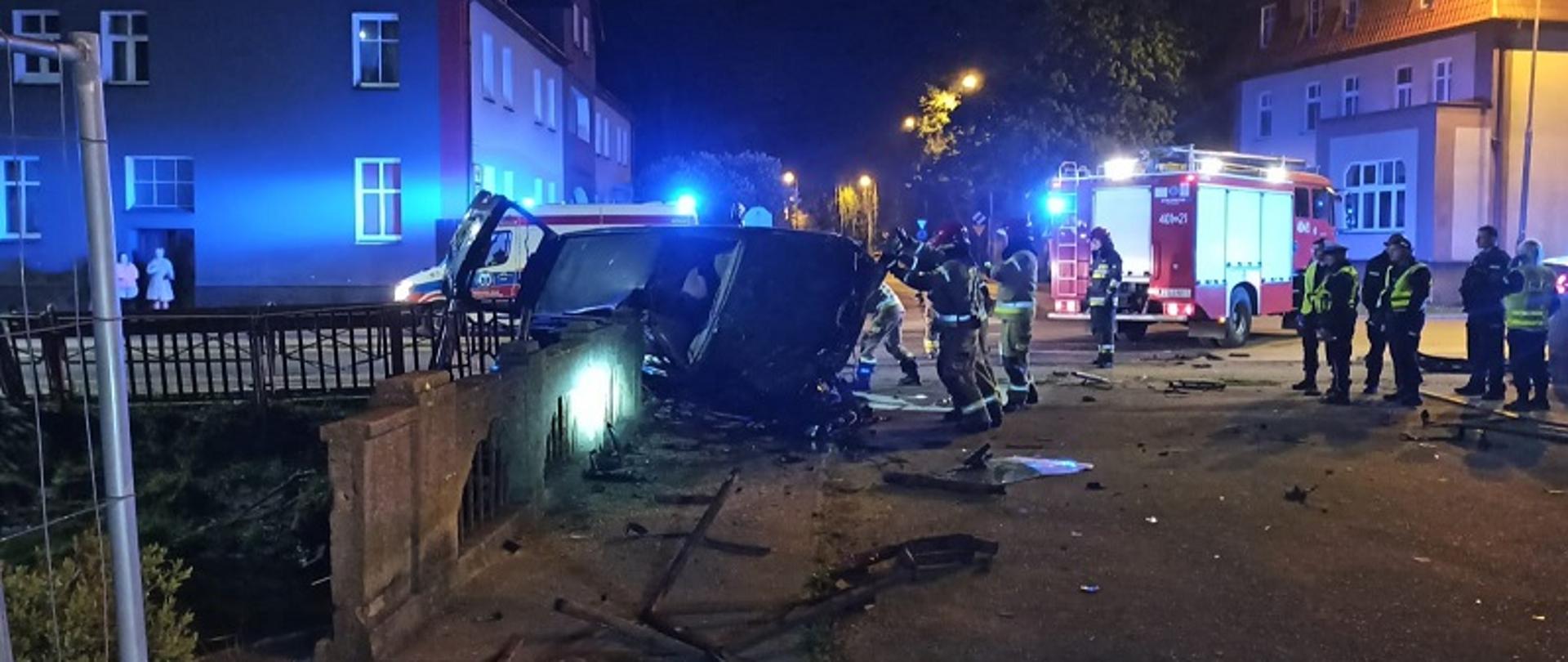 Zdjęcie wykonane nocą, przedstawia strażaków w ubraniach specjalnych zabezpieczających leżący na boku samochód osobowy. Pojazd częściowo oparty o konstrukcję mostu. W centralnym punkcie zdjęcia samochód strażacki, po prawej stronie funkcjonariusze Policji obserwujący działania strażaków. Przed wrakiem pojazdu elementy uszkodzonego ogrodzenia i fragmenty muru mostu
