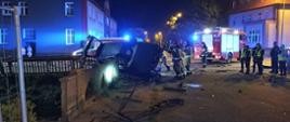 Zdjęcie wykonane nocą, przedstawia strażaków w ubraniach specjalnych zabezpieczających leżący na boku samochód osobowy. Pojazd częściowo oparty o konstrukcję mostu. W centralnym punkcie zdjęcia samochód strażacki, po prawej stronie funkcjonariusze Policji obserwujący działania strażaków. Przed wrakiem pojazdu elementy uszkodzonego ogrodzenia i fragmenty muru mostu