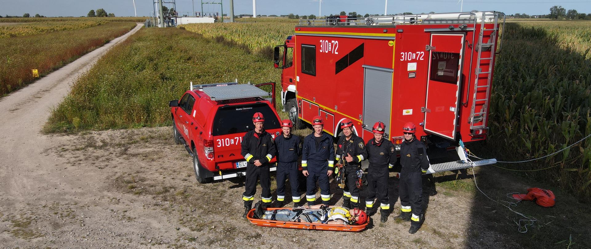 Sześciu ratowników pozuje do zdjęcia na tle wozów strażackich przed nimi manekin na desce ratowniczej.