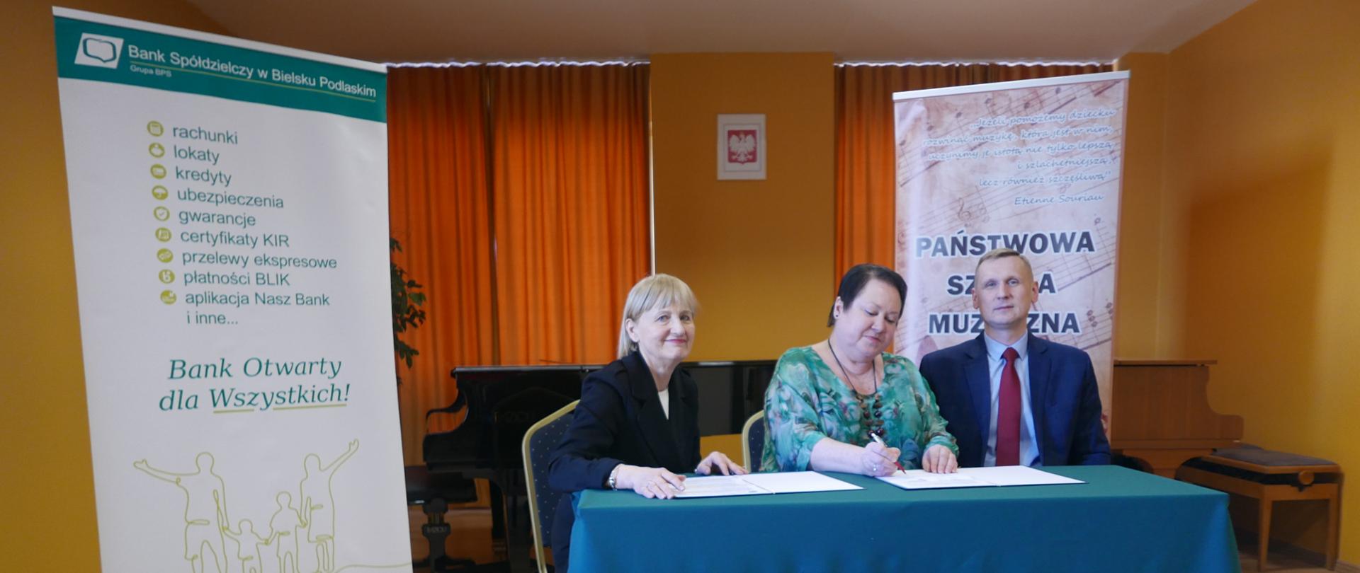Stolik przykryty ciemnozielonym materiałem. Na nim podpisywana jest umowa sponsorska. Przy stoliku trzy osoby-dwie panie i pan. Za nimi widoczne dwa rollupy z reklamą banku i szkoły muzycznej
