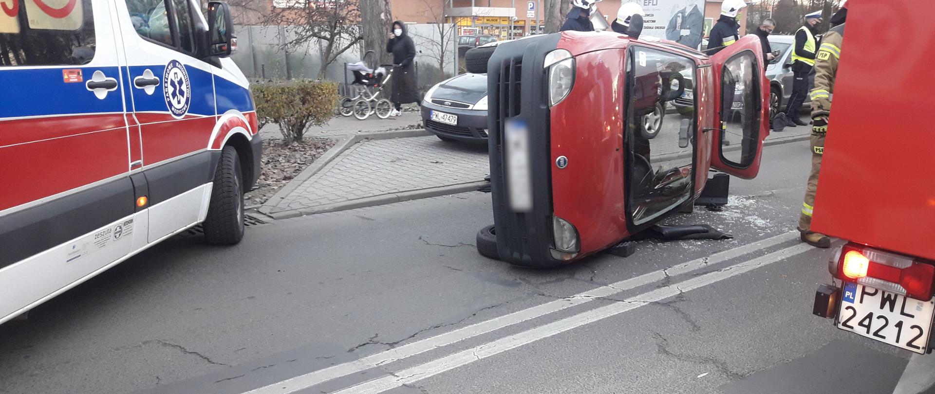 Na pierwszym planie leżący na boku samochód, w którym chwilę temu uwięziona była kobieta. Po lewej stronie karetka, po prawej samochód strażacki. W tle strażacy oraz policjanci w maseczkach.