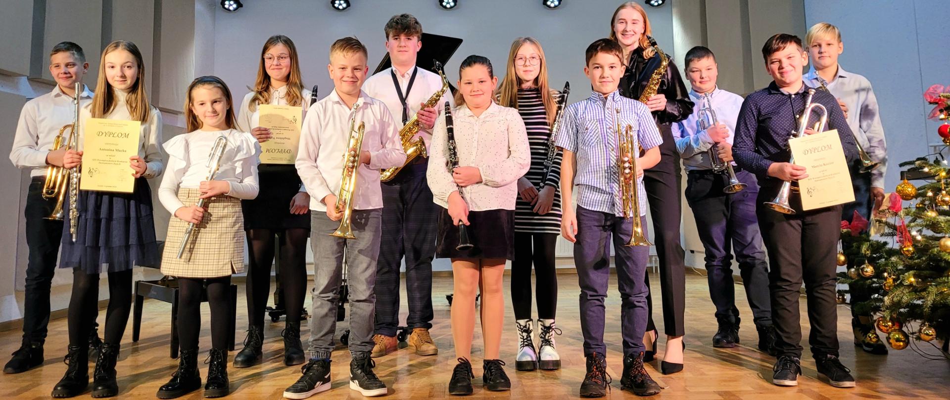 Zdjęcie przedstawia uśmiechniętych uczniów klas instrumentów dętych stojących na scenie szkoły. Dzieci trzymaja w rękach trąbki, klarnety, flety, saksofony oraz dyplomy. Za uczniami na środku sceny stoi czarny fortepian.