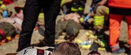 Fotografia przedstawia ratowników prowadzących czynności medyczne przy osobie poszkodowanej znajdującej się w pozycji leżącej. Jeden z ratowników wykonuje bezpośrednie czynności przy kończynie poszkodowanego, natomiast pozostali członkowie zespołu zabezpieczają pacjenta i wspierają prowadzoną procedurę.
W dalszej części kadru widoczni są kolejni poszkodowani oraz ratownicy realizujący równoległe działania. Taki sposób organizacji miejsca zdarzenia odzwierciedla warunki charakterystyczne dla zdarzeń o większej liczbie osób poszkodowanych, w których niezbędne jest prowadzenie jednoczesnych czynności ratowniczych wobec kilku osób oraz właściwe ustalanie kolejności udzielania pomocy.