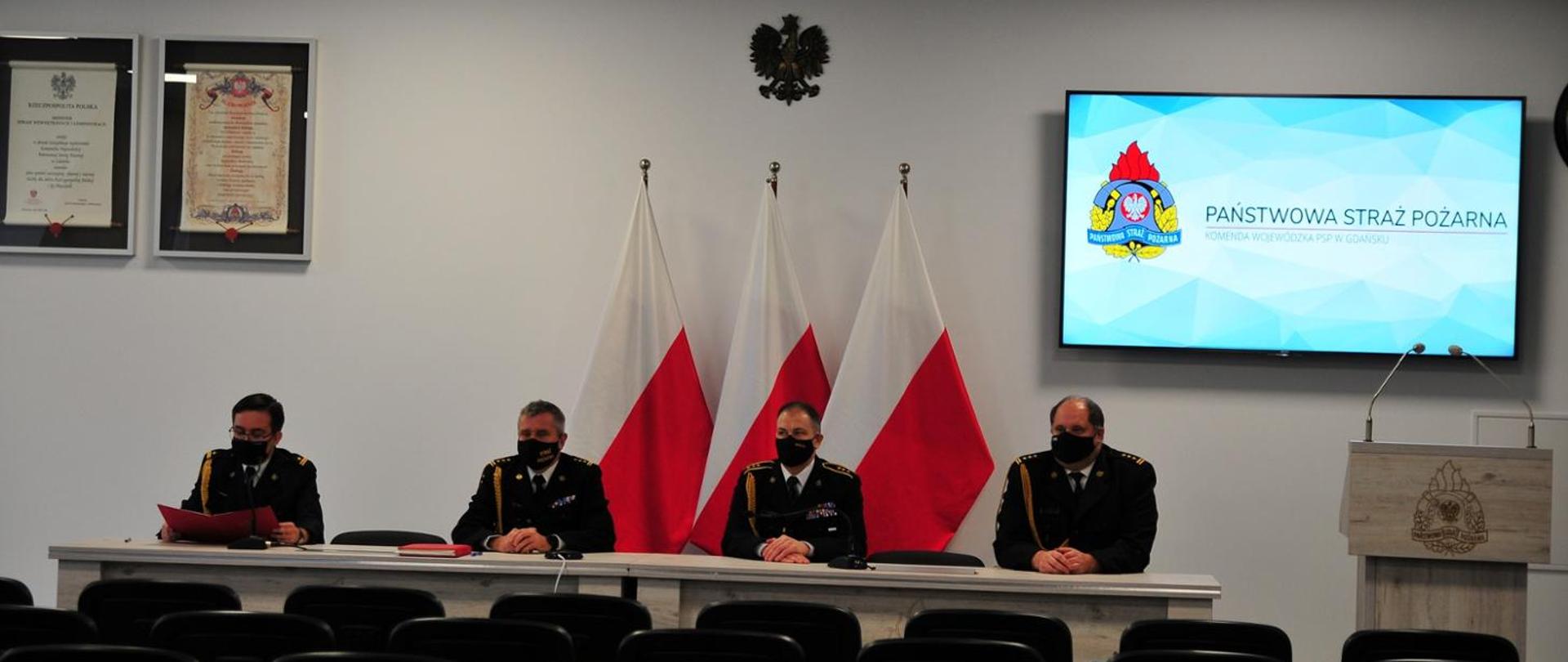 Czterech umundurowanych mężczyzn siedzi przy biurku na tle trzech polskich flag. Komendanci KW PSP w Gdańsku.