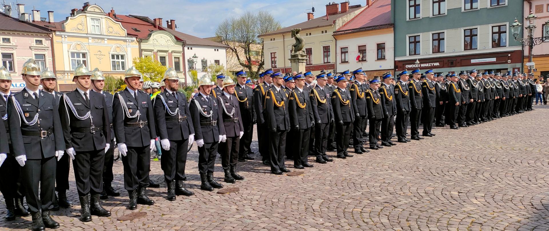Na zdjęciu w szeregu stoją strażacy w mundurach wyjściowych PSP i OSP. W tle widoczne zabytkowe kamienice rynku w Cieszynie.